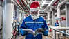 A photo of a worker in workwear, including protective clothing and a Santa Claus hat, standing in an ammonia plant. The worker is reading a magazine. The background contains pipes and equipment.