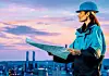 Frau in Arbeitskleidung mit Helm hält einen Plan in den Händen und schaut über eine Industrieanlage