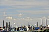 Blick über einen Industriepark mit Schornsteinen, Rohrleitungen und zahlreichen Anlagen unter leicht bewölktem Himmel.