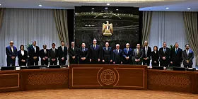 Delegation steht im Konferenzsaal vor großem Emblem hinter einem halbrunden Tisch.