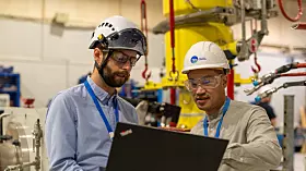 Zwei Ingenieure mit Helmen betrachten einen Laptop in einer Industrieanlage.