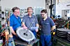 Drei Techniker betrachten ein großes Metallteil und studieren Pläne in einer Werkstatt.