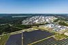 Luftbild zeigt Solarfeld vor Industrieareal, eingerahmt von Waldflächen.