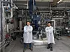 Zwei Personen in Schutzkitteln stehen vor einer großen Industrieanlage in einer Fabrikhalle.