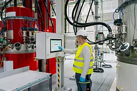 Arbeiter in Warnweste steht vor einem Bedienpanel in einer Industrieanlage mit Tanks und Rohren.