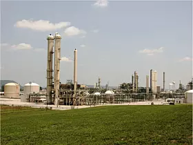Industrieanlage mit Türmen, Tanks und Rohrleitungen hinter einer grünen Wiese.