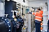 Techniker prüft Anlagen in einem Raum mit großen Pumpen, Rohren und technischen Geräten.