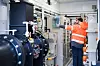 Techniker prüft Anlagen in einem Raum mit großen Pumpen, Rohren und technischen Geräten.