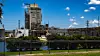 Industrieanlage mit hohem Turm, Leitungen und Grünstreifen an einer Straße unter blauem Himmel.