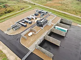 Luftaufnahme eines eingezäunten Industrieareals mit Gebäuden, Technikmodulen und Asphaltflächen.
