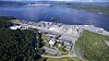 Luftaufnahme einer weitläufigen Industrieanlage an einer Küste mit Wasser und bewaldeten Hügeln.