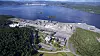 Luftaufnahme einer weitläufigen Industrieanlage an einer Küste mit Wasser und bewaldeten Hügeln.