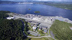 Luftaufnahme einer weitläufigen Industrieanlage an einer Küste mit Wasser und bewaldeten Hügeln.