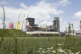 Große Baustelle mit Kränen, Rohbau und Wiese im Vordergrund unter blauem Himmel mit Wolken.