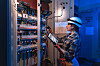 A professional electrician in safety helmet inspects an electrical cabinet using a tablet with technical diagrams inside an industrial facility. The scene reflects automation, smartfactory and modern digital maintenance.