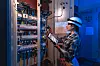 A professional electrician in safety helmet inspects an electrical cabinet using a tablet with technical diagrams inside an industrial facility. The scene reflects automation, smartfactory and modern digital maintenance.