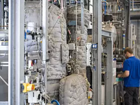 Arbeiter in einer Industrieanlage neben isolierten Rohren und technischen Anlagen.