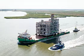 Großes Industrie-Modul auf einem Lastkahn, begleitet von Schleppern auf dem Wasser.