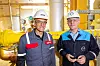 Zwei Arbeiter mit Schutzhelmen stehen vor gelben Rohren in einer Industrieanlage.