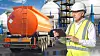 Industrial safety, Chemical plant. Engineer in hard hat inspects fuel tanker truck using tablet near storage tanks, performing logistics control and hazardous materials check.