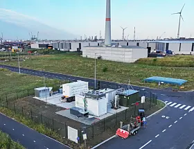 Eingezäunte Industrieanlage mit Containern, Straßen und Windrädern im Hintergrund nahe der Küste.
