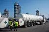 Großer zylindrischer Tank auf einem Tieflader, begleitet von Arbeitern in Warnwesten auf einem Industriegelände.