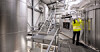 Arbeiter in Warnjacke prüft Geräte in einer hellen Industrieanlage mit Metalltanks und Treppen.