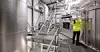 Arbeiter in Warnjacke prüft Geräte in einer hellen Industrieanlage mit Metalltanks und Treppen.
