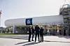 Vier Bauarbeiter in Helmen stehen vor einer großen Industrieanlage mit Gerüst und Rohrleitungen.