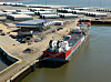 Frachtschiff an einem Industriehafen mit großen Rohrstapeln und Kränen am Ufer.