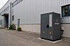 Grauer Stromspeicher steht vor einer Industriehalle an einer gepflasterten Zufahrt.