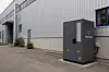 Grauer Stromspeicher steht vor einer Industriehalle an einer gepflasterten Zufahrt.