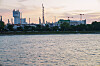 Industrieanlage mit hohem Prillturm am Ufer, im Vordergrund Wasser, im Hintergrund Kräne und Kamine.