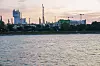 Industrieanlage mit hohem Prillturm am Ufer, im Vordergrund Wasser, im Hintergrund Kräne und Kamine.