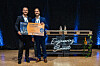 Zwei Männer zeigen eine orangefarbene „Startup of the Year 2024“-Tafel auf einer Bühne mit blauem Licht.
