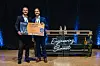 Zwei Männer zeigen eine orangefarbene „Startup of the Year 2024“-Tafel auf einer Bühne mit blauem Licht.