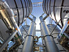 Blick nach oben auf große silberne Rohrleitungen und Metallstrukturen in einer Industrieanlage vor blauem Himmel.