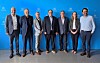 Sechs Personen stehen nebeneinander vor einer blauen Hintergrundwand mit Logos.