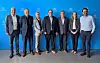 Sechs Personen stehen nebeneinander vor einer blauen Hintergrundwand mit Logos.